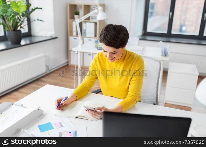 business, technology and user interface design concept - smiling ui designer with user interface templates and notebook using smartphone at office. smiling ui designer using smartphone at office