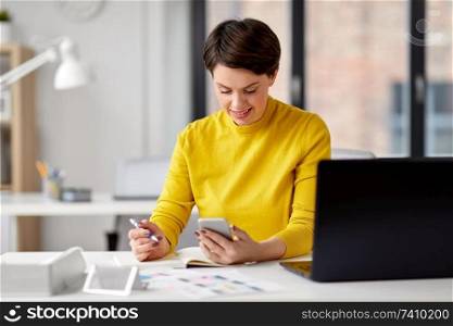 business, technology and user interface design concept - ui designer with notebook using smartphone at office. smiling ui designer using smartphone at office