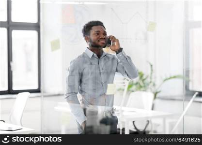 business, technology, communication and people concept - african american businessman calling on smartphone and looking at charts on office glass board. businessman calling on smartphone at office. businessman calling on smartphone at office