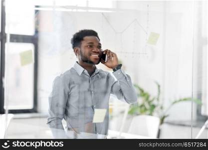business, technology, communication and people concept - african american businessman calling on smartphone and looking at charts on office glass board. businessman calling on smartphone at office. businessman calling on smartphone at office