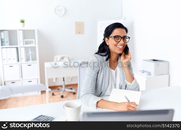 business, technology, communication and people concept - happy smiling businesswoman or secretary calling on smartphone and writing to notebook at office. businesswoman calling on smartphone at office