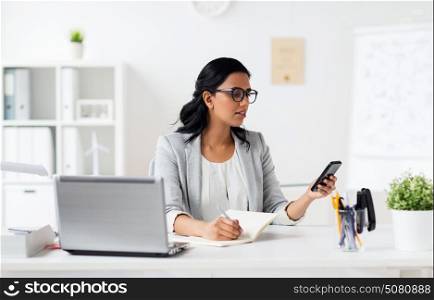 business, technology, communication and people concept - happy smiling businesswoman with smartphone and notebook writing at office. businesswoman with smartphone and laptop at office