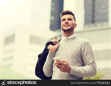 business, technology, communication and people concept - happy smiling young man in with bag and smartphone on city street. happy young man with smartphone and bag in city