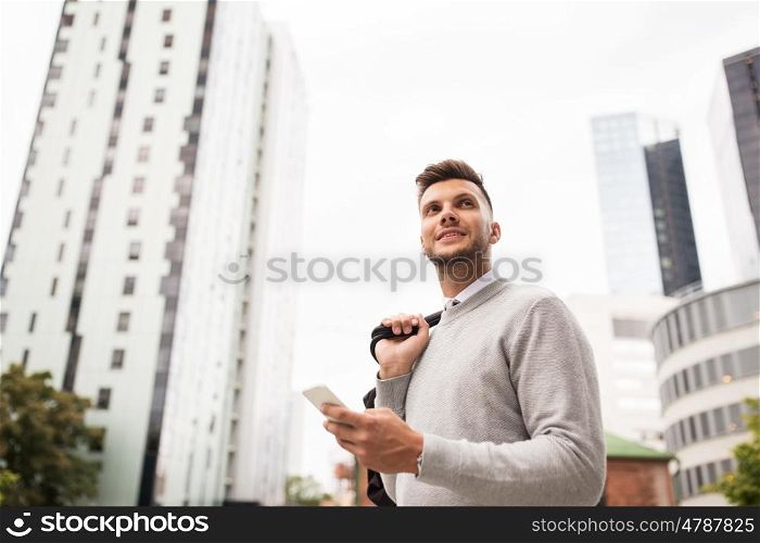 business, technology, communication and people concept - happy smiling young man with bag and smartphone on city street
