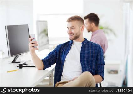 business, technology, communication and people concept - happy young creative man or student with computer at office texting on smartphone