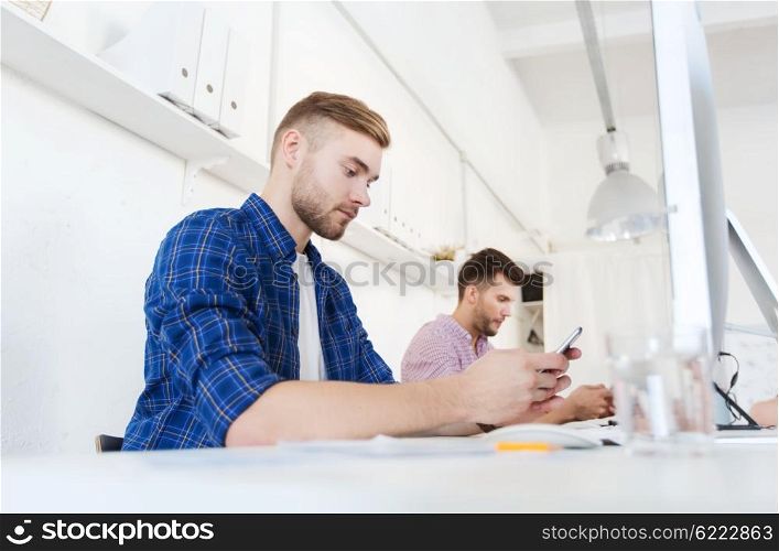business, technology, communication and people concept - young creative man or student with computer at office texting on smartphone