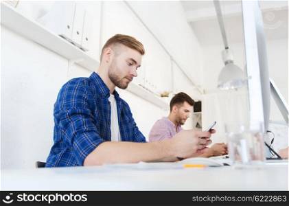 business, technology, communication and people concept - young creative man or student with computer at office texting on smartphone