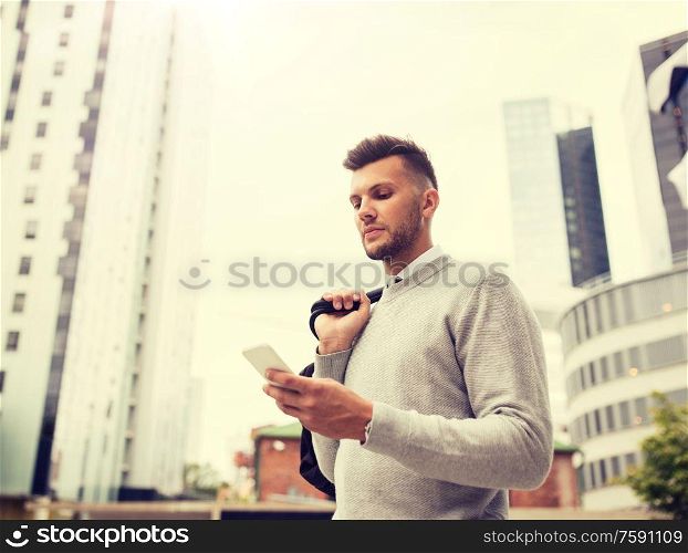 business, technology, communication and people concept - young man with bag texting on smartphone on city street. young man with bag texting on smartphone in city