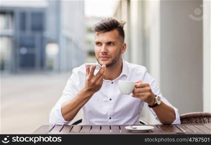 business, technology, communication and people concept - young man with coffee using voice command recorder on smartphone at city street cafe