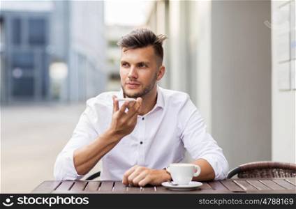 business, technology, communication and people concept - young man with coffee using voice command recorder on smartphone at city street cafe