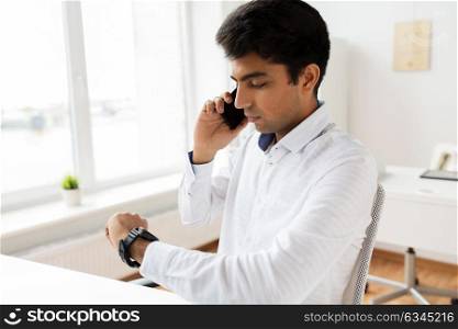 business, technology, communication and punctuality concept - close up of businessman calling on smartphone and looking at his wristwatch at office. businessman calling on smartphone at office