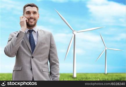 business, technology, innovation, energy saving and people concept - smiling businessman calling on smartphone over windmills and blue sky background
