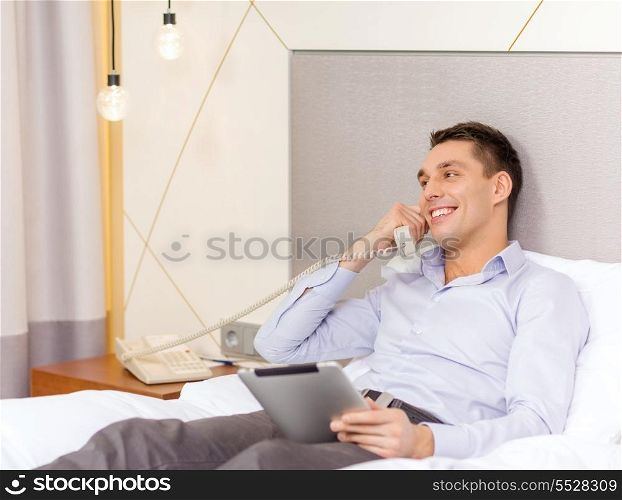 business, technology, internet and hotel concept - happy businessman with phone and tablet pc computer lying in hotel in bed