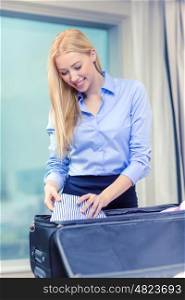business, technology, internet and hotel concept - happy businesswoman packing things in suitcase in hotel room