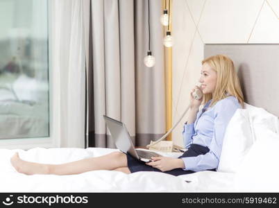 business, technology, internet and hotel concept - happy businesswoman with phone and laptop lying in hotel in bed