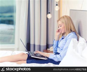 business, technology, internet and hotel concept - happy businesswoman with smartphone and laptop lying in hotel in bed
