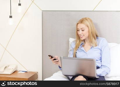 business, technology, internet and hotel concept - happy businesswoman with smartphone and laptop lying in hotel in bed