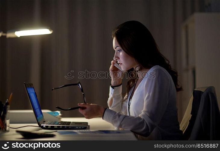 business, technology, overwork, deadline and people concept - woman with laptop calling on smartphone at night office. woman with laptop calling on smartphone at office. woman with laptop calling on smartphone at office