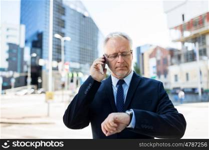 business, technology, time, punctuality and people concept - senior businessman calling on smartphone an looking at wristwatch or smart watch on his hand in city