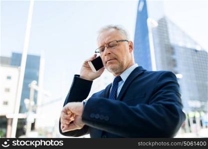 business, technology, time, punctuality and people concept - senior businessman calling on smartphone an looking at wristwatch or smart watch on his hand in city. senior businessman calling on smartphone in city