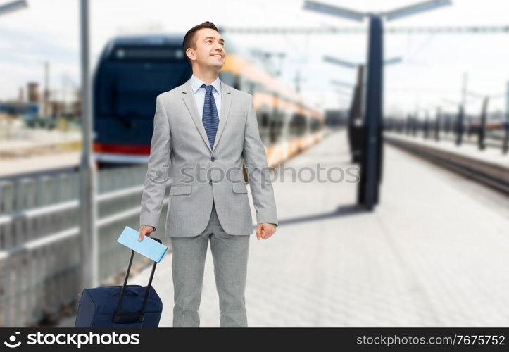 business trip and people concept - happy businessman in suit with travel bag and ticket over train on railway station on background. businessman with travel bag on railway station