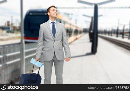 business trip and people concept - happy businessman in suit with travel bag and ticket over train on railway station on background. businessman with travel bag on railway station