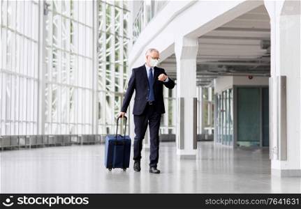 business trip, corporate and pandemic concept - senior businessman wearing face protective medical mask walking with travel bag along office building or airport and checking time. old businessman in mask with travel bag at airport