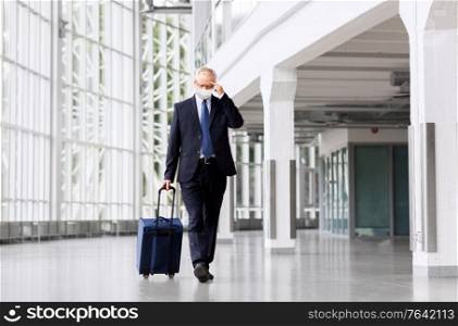business trip, corporate and pandemic concept - worried senior businessman in face protective medical mask walking with travel bag along office building or airport and thinking. old businessman in mask with travel bag at airport