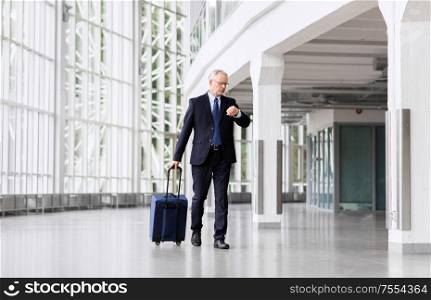 business trip, corporate and people concept - senior businessman walking with travel bag along office building or airport and checking time. senior businessman walking with travel bag