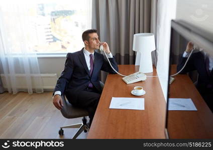 business trip, people and communication concept - businessman with coffee and papers calling on desk phone at hotel room. businessman calling on desk phone at hotel room