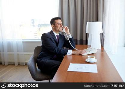 business trip, people and communication concept - happy smiling businessman with coffee and papers calling on desk phone at hotel room. businessman calling on desk phone at hotel room