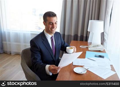 business trip, people and paperwork concept - happy smiling businessman with papers drinking coffee at hotel room. businessman with papers drinking coffee at hotel