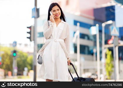 business trip, people and tourism concept - happy young asian woman with travel bag calling on smartphone on city street. woman with travel bag calling on cellphone in city