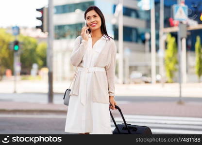 business trip, people and tourism concept - happy young asian woman with travel bag calling on smartphone on city street. woman with travel bag calling on cellphone in city