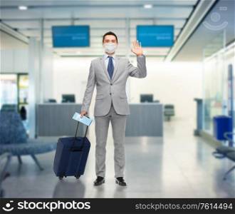 business trip, traveling and health care concept - happy businessman in suit wearing face protective medical mask with travel bag and air ticket waving hand over airport background. businessman in mask with travel bag at airport