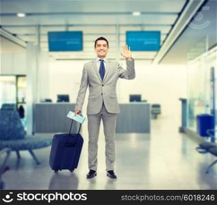 business trip, traveling, luggage and people concept - happy businessman in suit with travel bag and air ticket waving hand over airport background