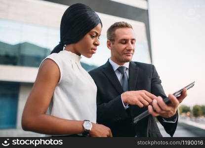 Business woman and businessman looks on laptop screen, outdoors meeting of partners, modern office building on background, partnership negotiations during the lunch break. Successful businesspeople