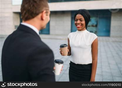 Business woman and businessman with coffee cups, outdoors meeting of partners, modern office building on background, partnership negotiations during the lunch break. Successful businesspeople