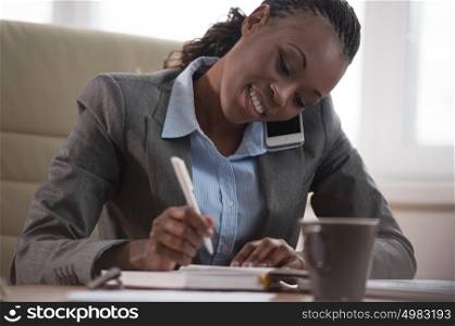 Business woman calling phone and writing something in her schedule book and papers at office workplace