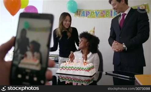 Business woman celebrating her birthday and doing a party with colleagues in her office. A friend records a video on mobile phone of her blowing out clandles on cake. Medium shot