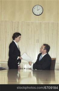 Business woman hands over papers to business man who is sitting at the head of the conference table