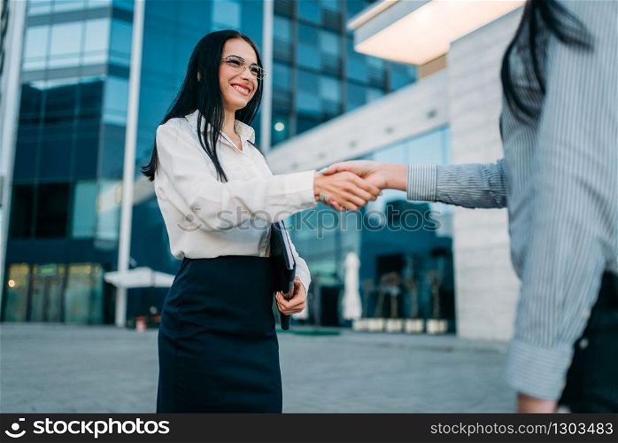 Business woman in glasses and white blouse, handshake with partner outdoor. Modern building, financial center. Successful female businessperson