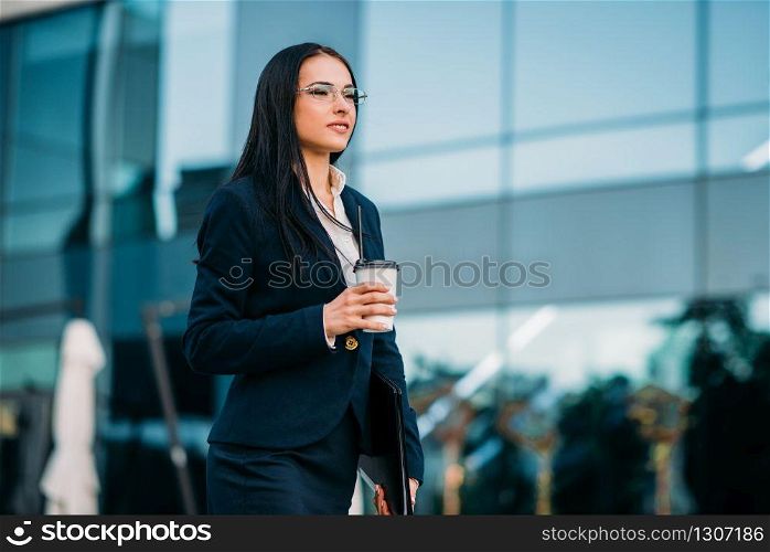 Business woman in glasses with coffee in hands, against skyscraper. Modern building, financial center, cityscape. Successful female businessperson