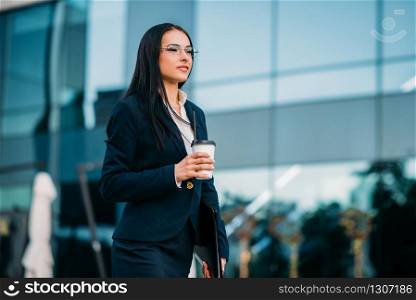 Business woman in glasses with coffee in hands, against skyscraper. Modern building, financial center, cityscape. Successful female businessperson