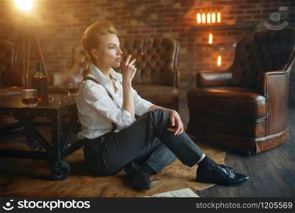 Business woman in strict clothes relax on the floor with whiskey and cigar, retro fashion, gangster style, female macho. Vintage lady in office with brick walls. Business woman relax with whiskey and cigar