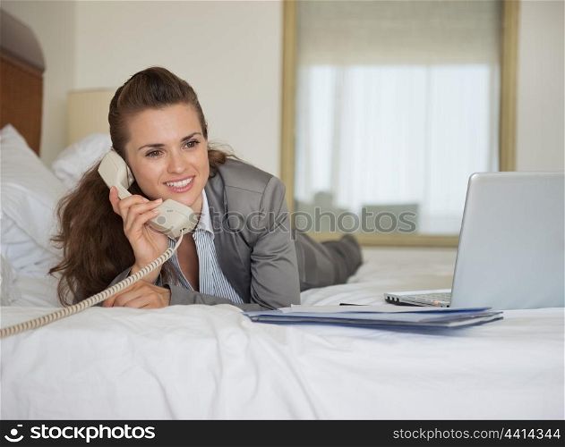 Business woman laying in bed with laptop and talking phone in hotel room