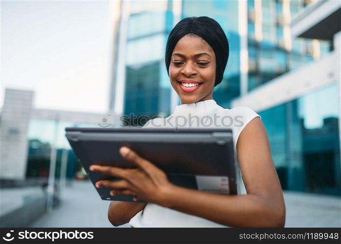 Business woman looks on laptop screen in front of office building. Smiling black businesswoman in white blouse works on pc outdoors