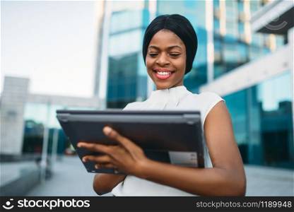 Business woman looks on laptop screen in front of office building. Smiling black businesswoman in white blouse works on pc outdoors