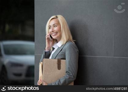 Business woman portrait outdoors talking by phone and holding documents with modern building as background.