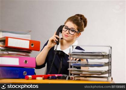 Business woman sitting working at desk full off documents in binders talking with someone on phone. Business woman in office talking on phone